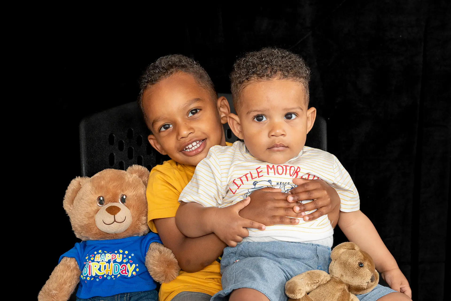 Older brother and younger, sat smiling for a photograph, with teddy bear with happy birthday t-shirt on