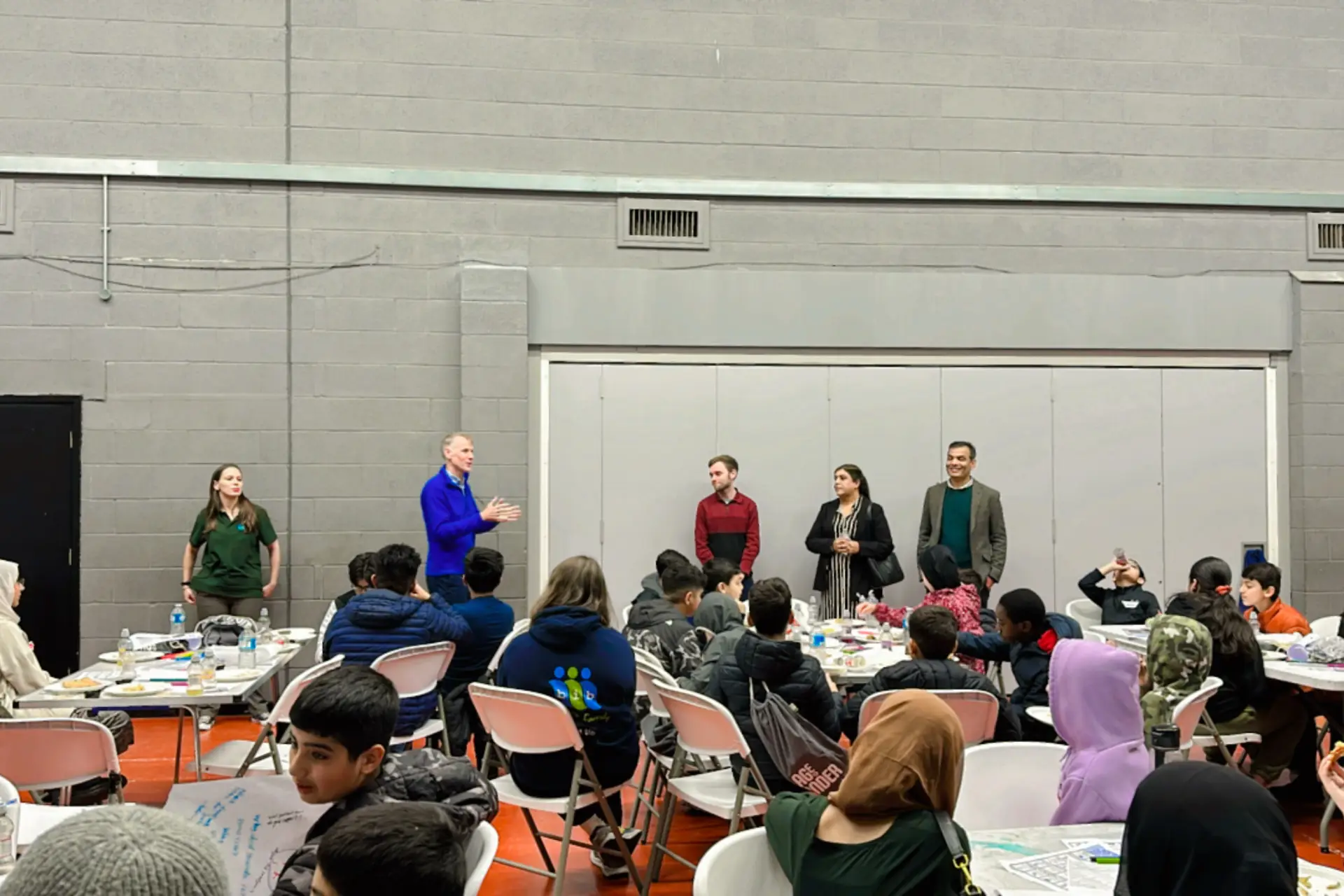 Children in a school sat around tables, with teachers and volunteers stood up presenting